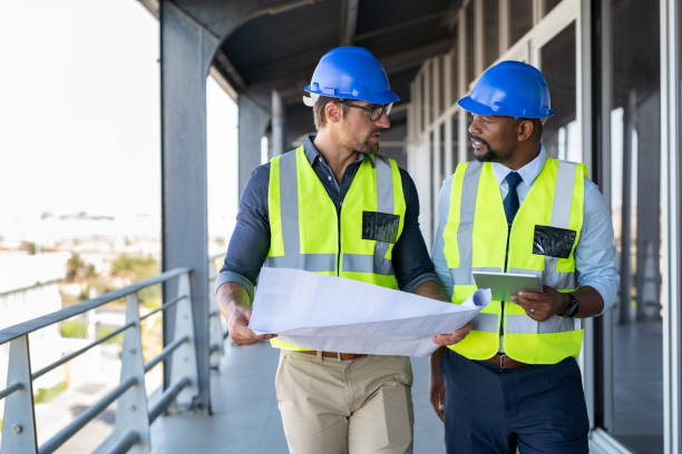 Metalworker and engineer meeting at construction site. Multiethnic architects in construction site with safety equipment walking and discussing. Architect and engineer working together while visiting a new building.